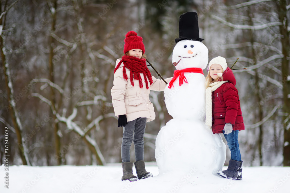 Two adorable little girls building a snowman together in beautiful winter park. Cute sisters playing in a snow.