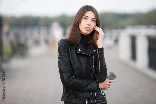 Close up emotional portrait of a young pretty brunette woman posing full length outdoors city park wearing black leather coat holding smartphone