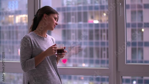 Young woman standing by window drinks coffee HD slow-motion video. Girl looking at morning or evening city landscape at home flat apartment photo