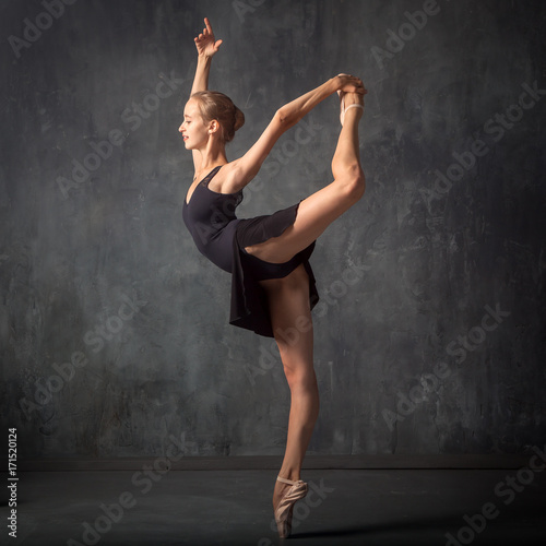 A young beautiful ballerina in a black dress, white pantyhose and pointe shoes beautifully poses and dances ballet in an image in a dark dance stage