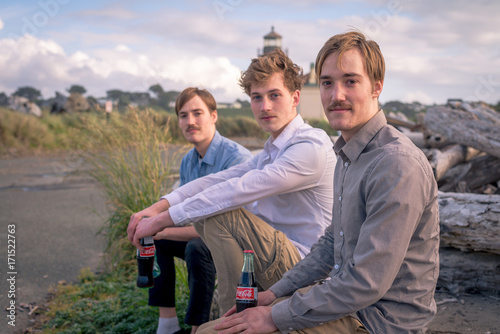 Portrait of three friends sitting on a log outdoors by a lighthouse     © mdurson