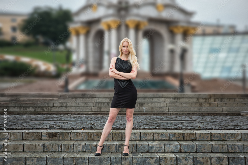 Beautiful blonde young woman in black dress walking dawn stairs outside in Kyiv. Rainy day