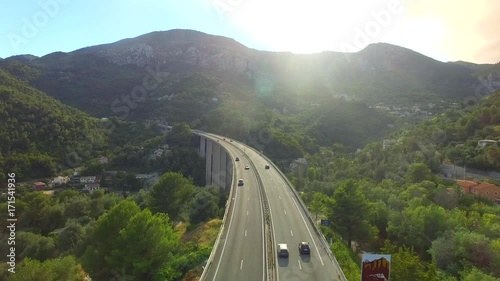 Cars move at highway bridge road in mountains above valley village morning aerial 4k. Fly above traffic on bypass over hill town in Europe sun shine. Motorway vehicles in scenic Italian Alps landscape