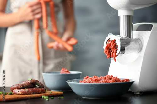 Meat grinder with fresh forcemeat and woman holding sausages in kitchen photo