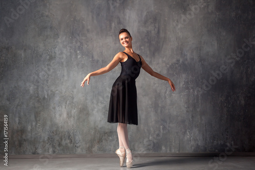 Young beautiful ballet dancer with gathered hair in a bun in a black dress, pointe shoes smiling and beautifully dancing ballet in a dark dance studio