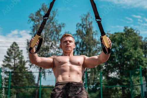 Male athlete is an excellent trx training, fresh air nature in summer, feel your strength and balance, motivation, tanned skin. Exercise of muscle of the chest. A sunny day. Stamina of a man. Against