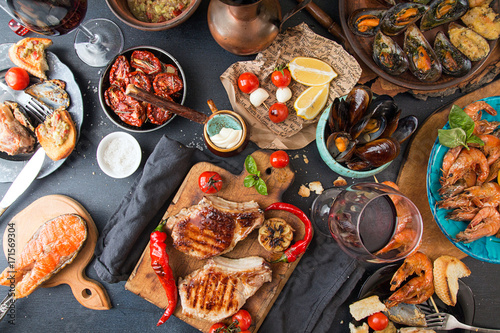 Overhead of dinner table. Assorted grilled barbecue meat and seafood with vegetable. Pork grilled steaks, salmon trout, mussels, shrimps, dried tomato, cherry tomato, chilii pepper, glass of vine.