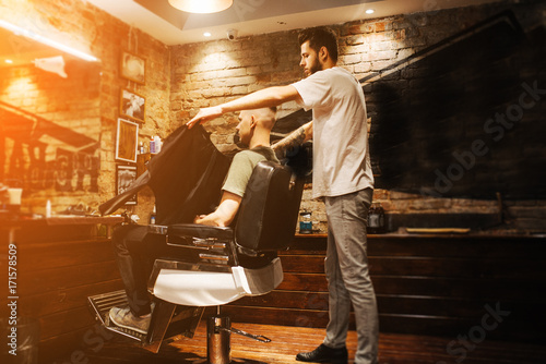 Grooming of real man. Side view of young man sitting in chair at barbershop. The hairdresser throws a cloak over the client. Preparing for a haircut. Underground. Grunge style with a little noise.