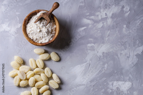 Raw uncooked potato gnocchi with olive wood bowl of flour, grated parmesan cheese over gray concrete background. Top view with copy space. Home cooking.