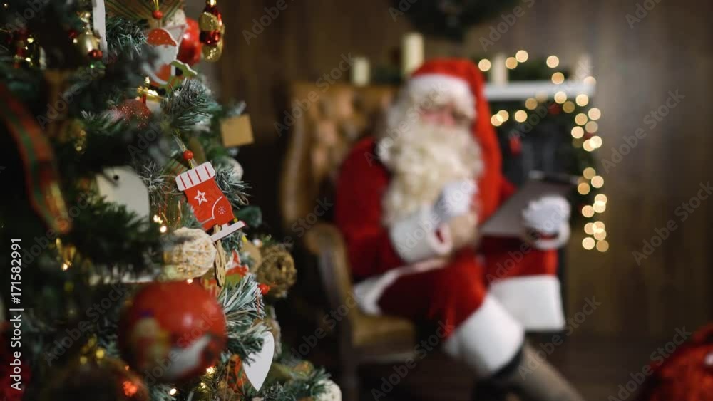 Blurred Santa Claus working with tablet PC, focus on christmas green tree with lights and toys and decorated fireplace. dolly slider shot. DigitalPad Blurred Santa Claus working with tablet PC, focus on christmas green tree with lights and toys and decorated fireplace. dolly slider shot. DigitalPad