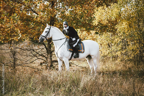 Weird woman in a bird (penguin) rubber mask wearing a hat, riding a white horse in the autumn forest © anastasianess