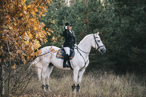 Young attractive brunette riding her white horse, wearing equestrial uniform in the autumn forest