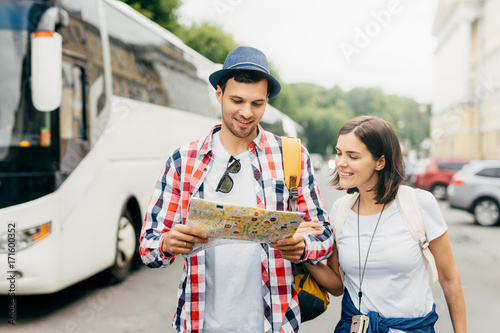 Cheerful guy in hat and shirt, holding city map, looking with smile in it, standing near his female friend, walking in city, exploring new entertainments. Male and female tourists with city guide. photo