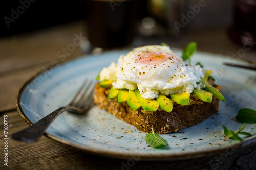 Black bread toast with fried benedict egg, avocado and salmon spread in blue plate with coffee in background, wooden shabby table with coffee pot, glass and books stack
