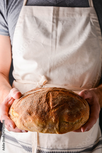 Man Wearing Apron Holding Freshly Baked Loaf Of Bread.