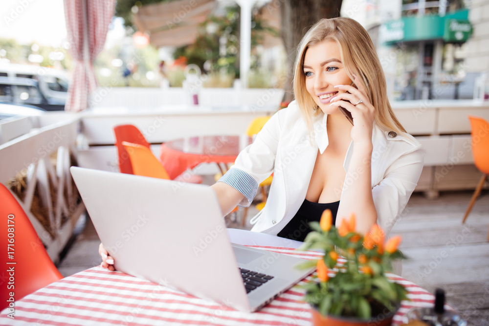 Young attractive girl talking on mobile phone and smiling while sitting alone in coffee shop during free time and working on tablet computer. Happy female having rest in cafe.