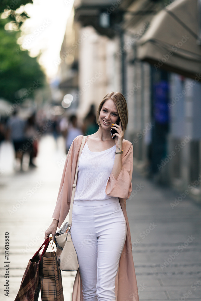 Fototapeta premium Beautiful and fashionable young woman with shopping bags standing on city street and looking at shopfront