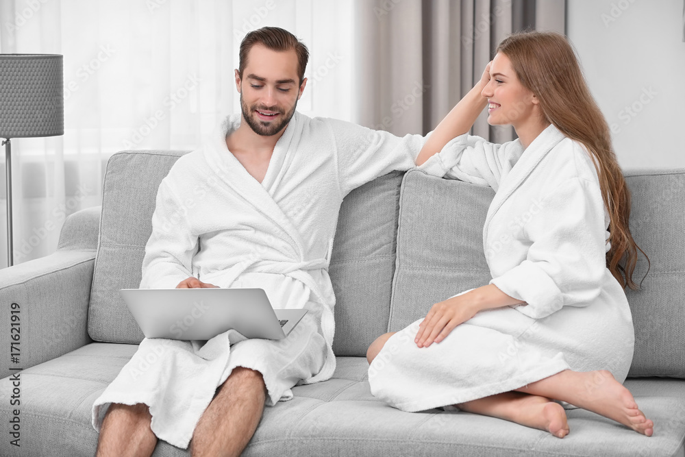 Young loving couple in bathrobes with laptop sitting on sofa at home Young loving couple in bathrobes with laptop sitting on sofa at home