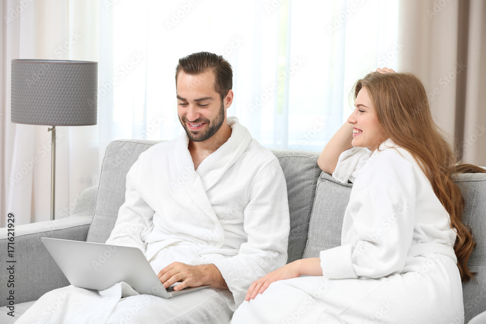 Young loving couple in bathrobes with laptop sitting on sofa at home Young loving couple in bathrobes with laptop sitting on sofa at home