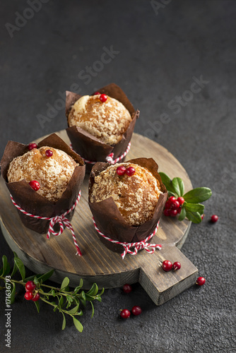 A festive breakfast. Fragrant fruitcakes in brown paper forms with berries of lingonberry on wooden background with berries, New Year red balls. Selective focus
