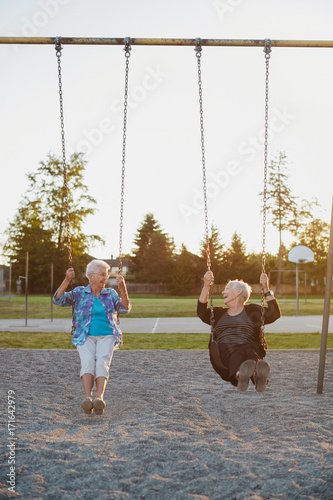 Two happy, senior caucasian friends swinging together on swingset at sunset