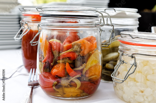 Glass preserving jars of pickled chillies, onions and gherkins on white table.