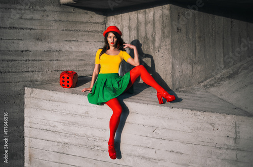 Fashion portrait of a young girl in a colorful clothes with a red fashionable handbag. Grey geometric abstract background