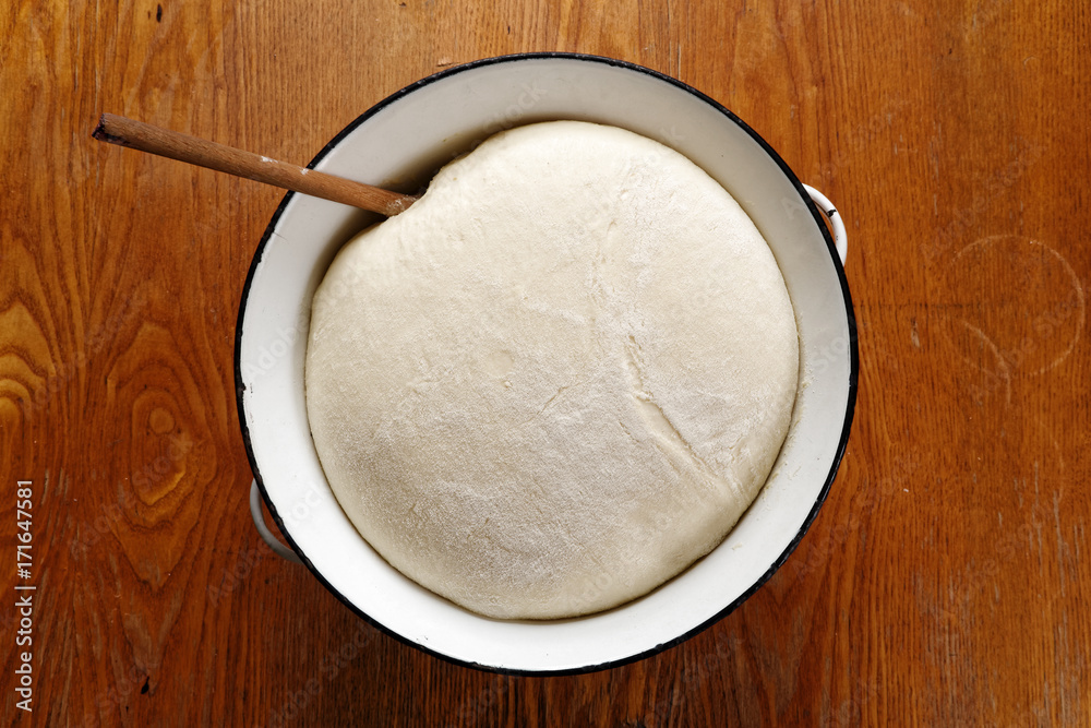Leavened yeast dough with wooden spoon in white enamel metal bowl on wooden table from above.