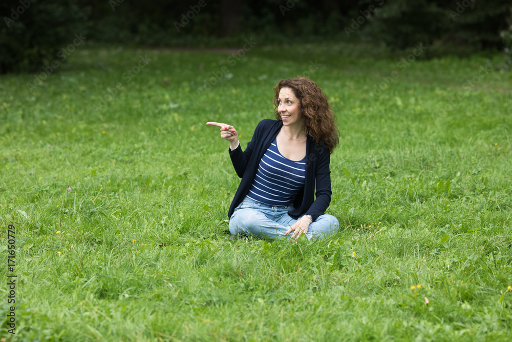 Fototapeta premium A young woman is sitting on the grass and pointing at something with her finger