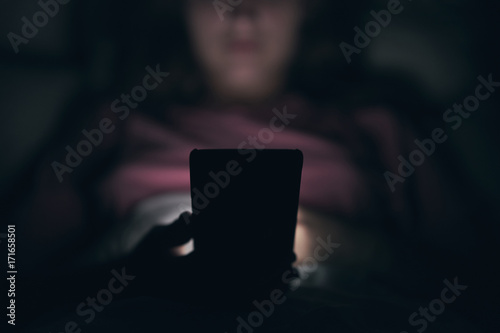 Close-up. A young girl is typing on a smartphone. Lying in bed. Blurred dark background. At night