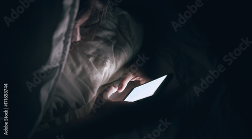 the girl is using the phone. Lying in bed. Dark blurred background