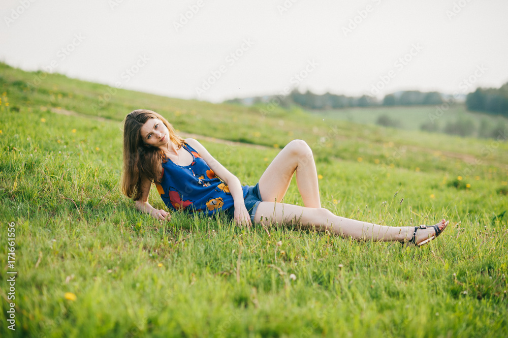 Young skinny girl lying on grass on hillside at nature outdoor. Lonely  beautiful brunette teen with naked long legs looking at camera in  countryside wild terrain. Soft warm evening autumn weather. Stock