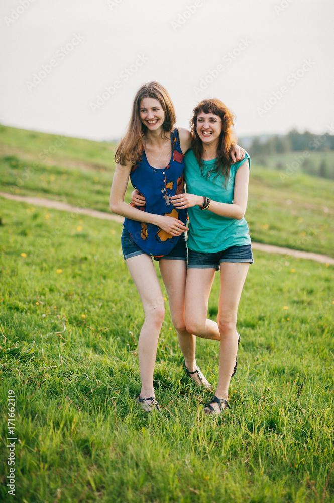 Fototapeta premium Two young lovely girls with long hair and naked legs spending time at nature outdoor. Women friendship. Weekend with beloved friend. Expressive emotion. Laughing and smiling teenagers. Youth lifestyle