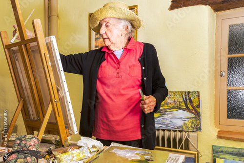Senior woman, wearing a  sun hat, painting at her easel, in her studio in France.