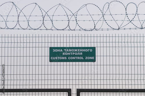 Customs control zone sign (written in Russian and English) on a fence with barbed wire with the gray skies in the background. A concept of Russian sanctions, bans, border and trade