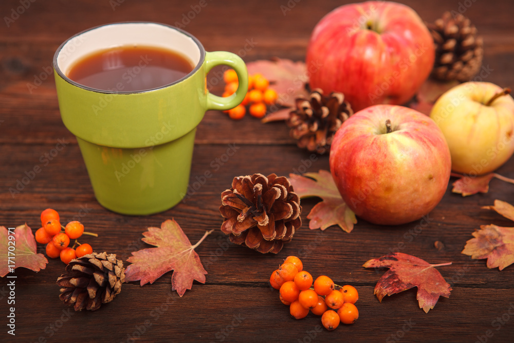 Autumn concept, fallen red-yellow leaves with apples, pine cones, rowan and a cup of tea on a wooden table. Thanksgiving Day.