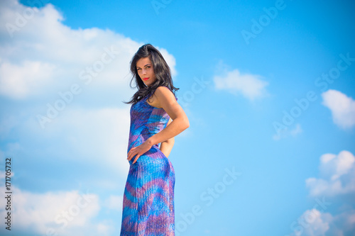 Woman with brunette curly hair in blue-purple long dress near sand dunes against sky background. Sexy stylish fashionable thin lady posing in desert
