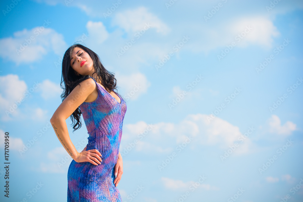 Woman with brunette curly hair in blue-purple long dress near sand dunes against sky background. Sexy stylish fashionable thin lady posing in desert Woman with brunette curly hair in blue-purple long dress near sand dunes against sky background. Sexy stylish fashionable thin lady posing in desert