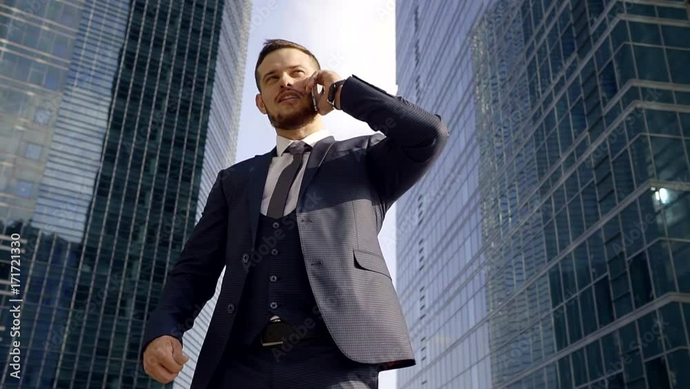 young manager is wearing three-piece suit is talking by phone, standing in background skyscrapers