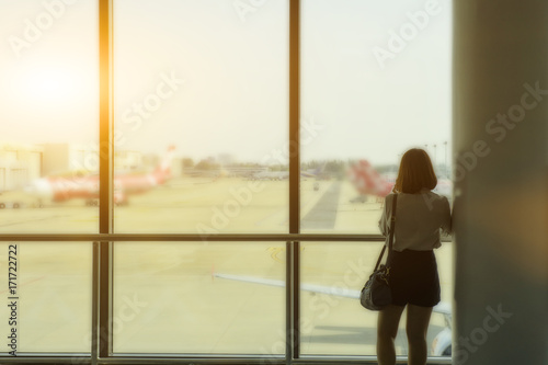 Short hair asian lady girl standing againt glass wall looking at airplane in airport while waiting for flight.