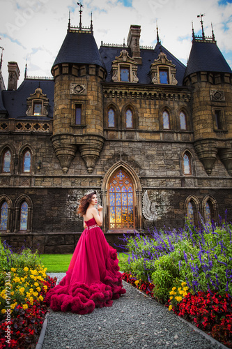 A beautiful woman, a queen in a burgundy lavish dress, walks along a flowering garden. Ancient castle on the background. photo