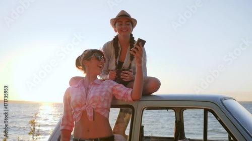 photo at mobile of Beautiful Friends near automobile on shore river, Lovely girls Photographed near vehicle on Embankment, girlfriends Makes Selfphoto at Car, backlighting blue sky, photo