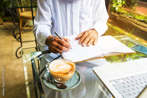 Arab muslim business man ware white traditional clothing and write on paper ,On table have laptop smartphone and cup of coffee