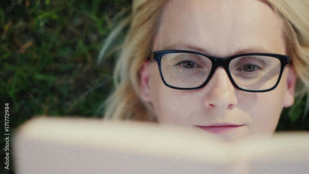 Portrait of a young woman in glasses, lying on a lawn in the park and reading a book. Close-up Portrait of a young woman in glasses, lying on a lawn in the park and reading a book. Close-up