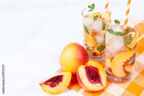 Peach lemonade with ice and mint leaves. Homemade lemonade of ripe nectarine with white and orange ripe. Two glasses of peach tea. Refreshing summer drink on a white wooden background.