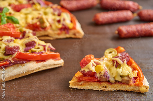 Tasty piece of pizza with salami, tomatoes, bell pepper, onion, green olives, corn, cheese and spices. On blurred background sliced pizza and little sausages salami on brown wooden table close up