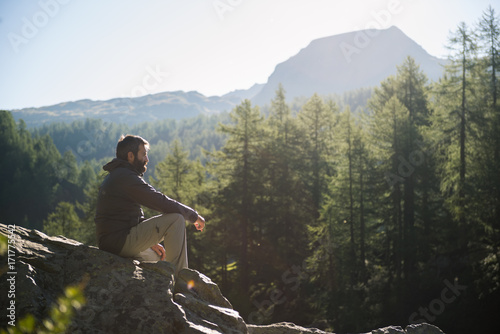 Young adult active man sitting on top rock looking at panorama in sunny spring summer morning on mountain outdoor.