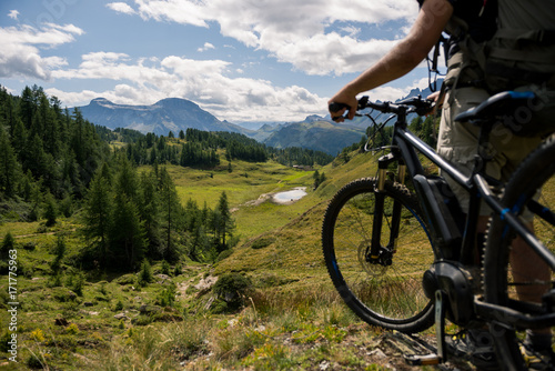 Young adult active man on mountain wearing bike helmet and backpack looking at scenic panorama holding electric bike in sunny summer day outdoor.
