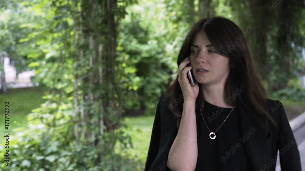 Young and attractive businesswoman wearing black clothes is talking on her smartphone and walking in a park. Handheld real time establishing shot Young and attractive businesswoman wearing black clothes is talking on her smartphone and walking in a park. Handheld real time establishing shot