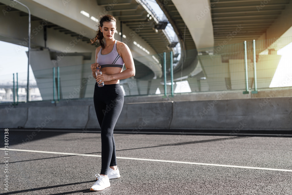 Full length shot of young sporty woman standing under a bridge, relaxing after workout